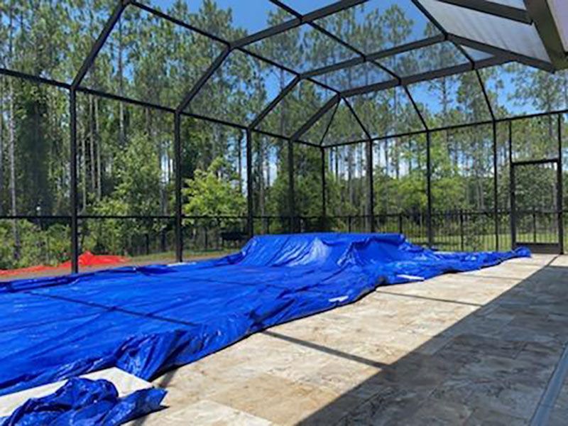 Blue tarp covers a screened pool enclosure floor beside a concrete patio and trees.