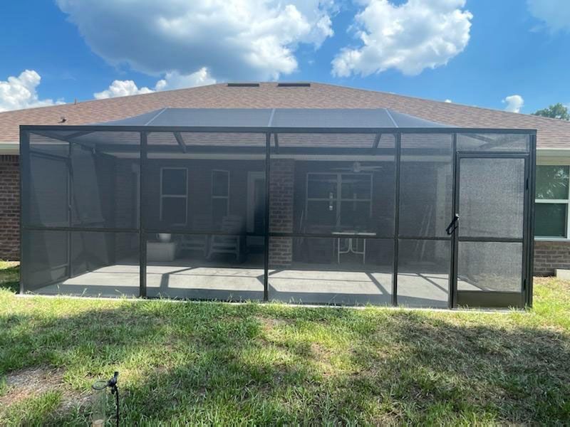 Screened patio enclosure in a backyard with a roof and black mesh panels under a blue sky.