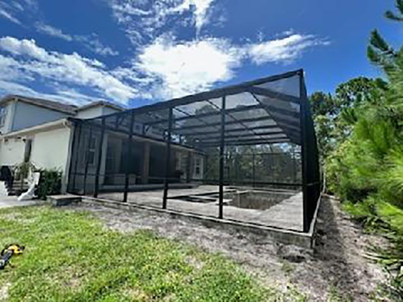 Screened patio enclosure beside a house, with black metal framing and a concrete floor under a blue sky.