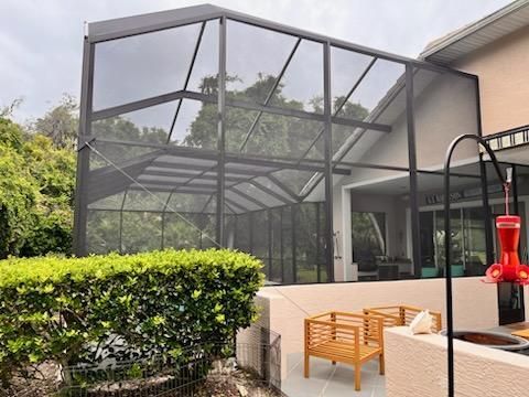 Screened backyard patio with wicker seating beside a beige house and hedge-lined pool area