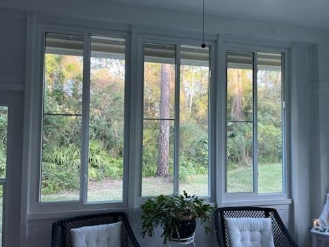 Large living room windows with white trim, overlooking a green garden, with two chairs and a potted plant below.