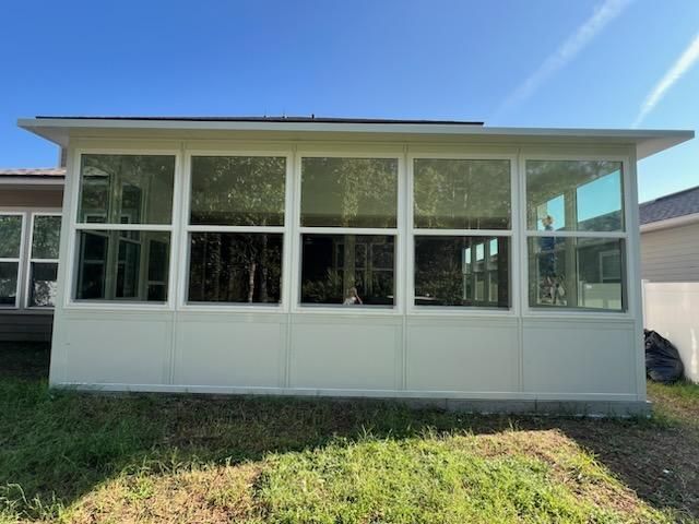 White sunroom/screened porch with large windows and a flat roof in a grassy yard.