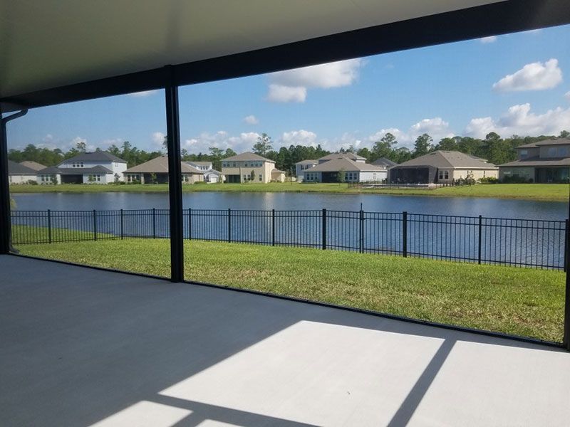 Covered patio overlooking a fenced backyard and lake with houses across the water under a blue sky.