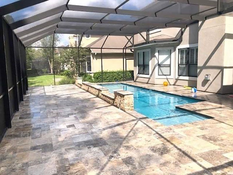 Covered patio with a swimming pool, stone tile floor, and screened enclosure beside a house.