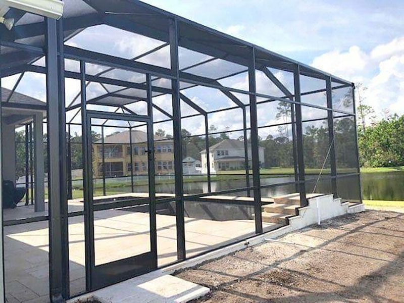 Black metal screened pool enclosure beside a backyard patio and water.