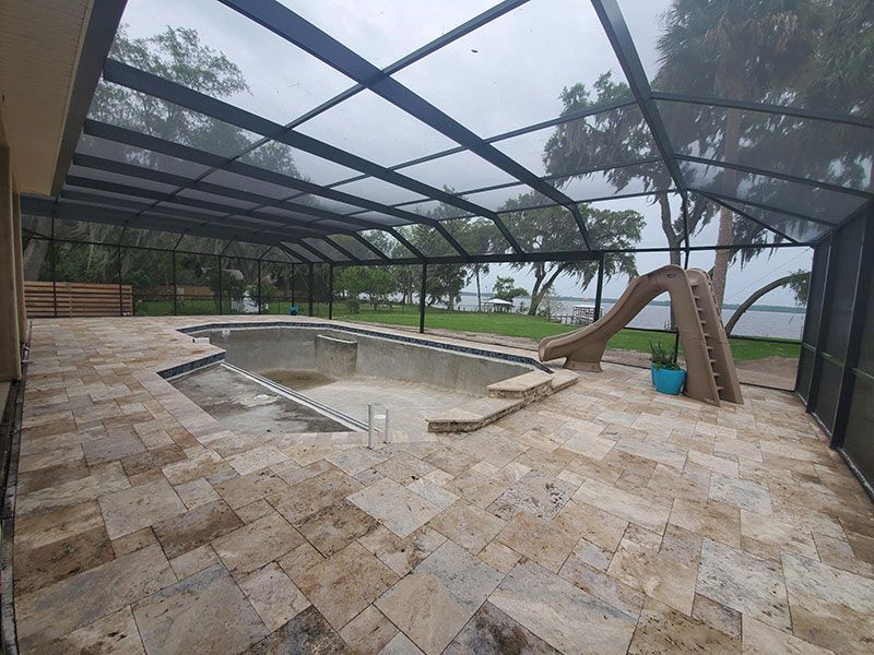 Screened patio with empty pool, stone deck, and a children’s slide beside a lake