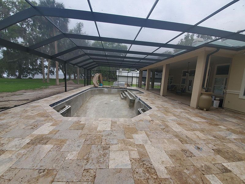 Empty rectangular pool beside a covered patio with stone decking and a black screen enclosure