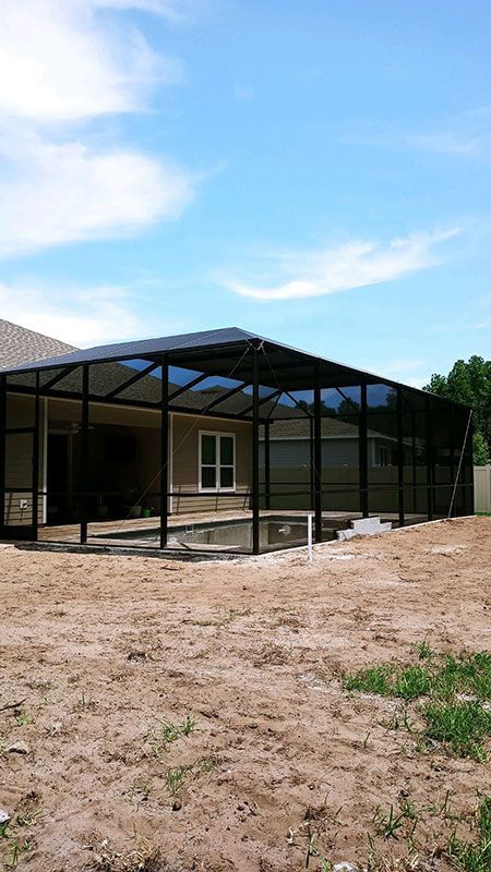 Black metal screened patio enclosure beside a house on a dirt lot under a blue sky