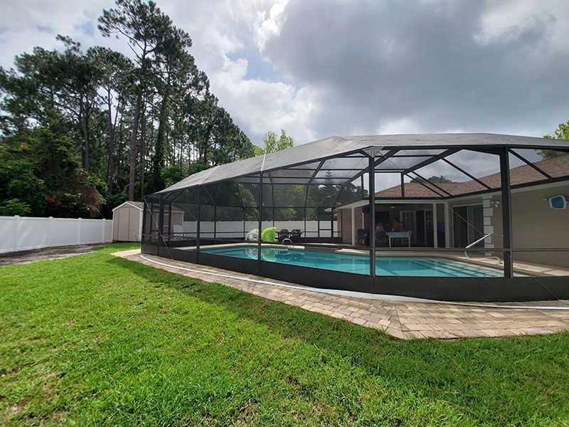 Backyard pool with screened enclosure beside a house, surrounded by grass and trees under cloudy skies