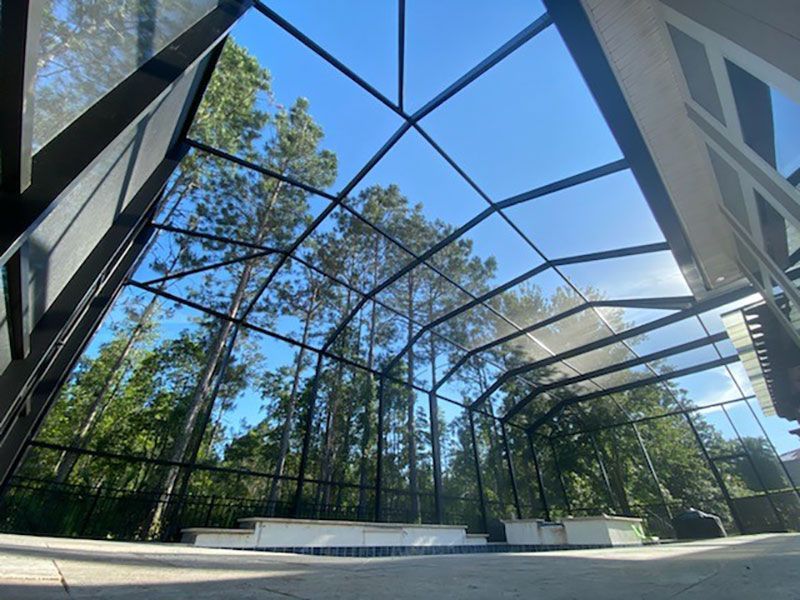 Outdoor tennis court with a black net canopy, white benches, and tall trees under a blue sky