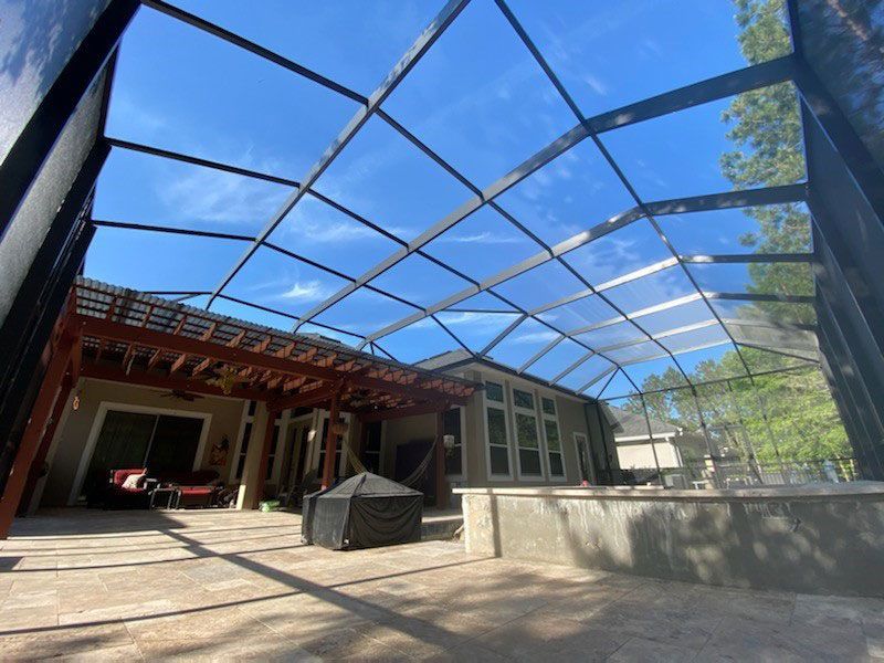 Open-air patio with blue screened roof, concrete floor, and red-roofed structure in daylight