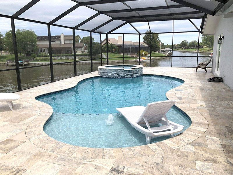 Screened lanai with a blue freeform pool, spa, and white lounge chair overlooking water.