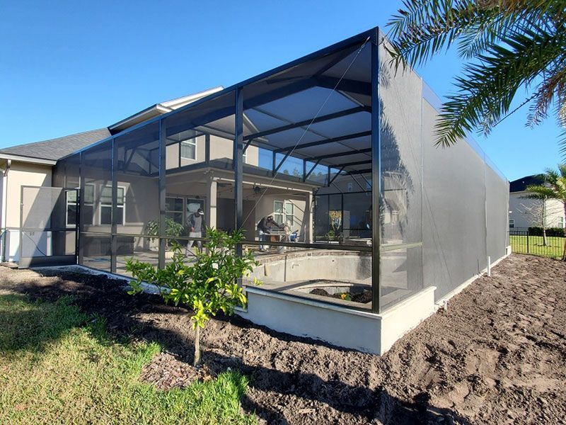 Screened patio enclosure attached to a house, with mesh walls, dark roof framing, and sandy yard.