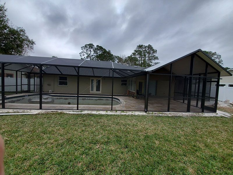 Covered fenced pool beside a beige house with screened enclosure and green lawn under cloudy sky