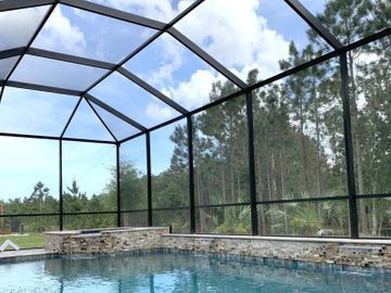 Screened backyard pool with stone coping, black frame enclosure, and trees beyond.