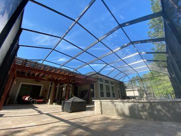 Covered patio with a metal screen roof, concrete floor, and a house opening onto a sunny backyard