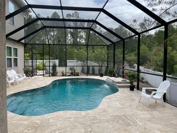 Screened-in backyard pool with white chairs and stone patio under a black enclosure