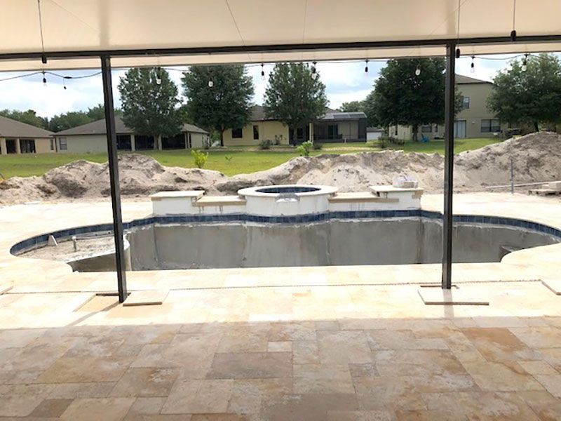 Empty backyard swimming pool under construction with tan tile patio and dirt piles in the background