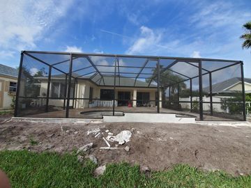 Screened patio enclosure under construction behind a house, with dirt foreground and blue sky.