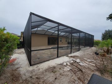 Black metal screened patio enclosure attached to a beige house in a dirt yard.