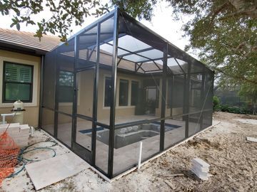 Black-framed screened patio enclosure beside a house, with stone flooring and a small hot tub inside.