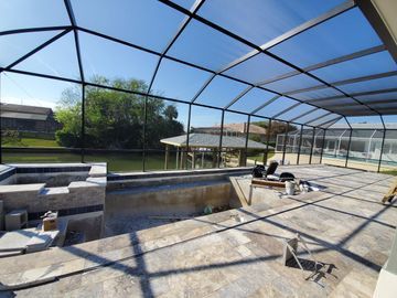 Screened pool enclosure with a partially finished pool deck and patio furniture in sunlight