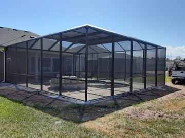 Black screened-in patio enclosure on a grassy backyard beside a house, under a blue sky
