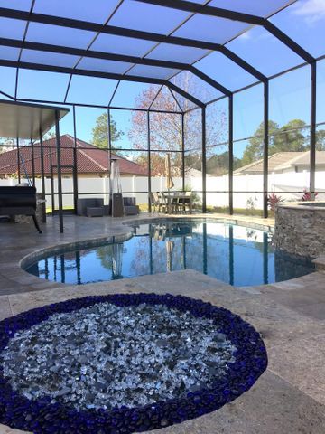 Screened-in backyard pool with a stone patio and circular blue pebble fire pit at sunset