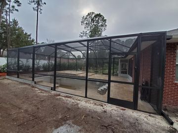 Black mesh enclosure around a concrete pool patio beside a brick house and wooded yard