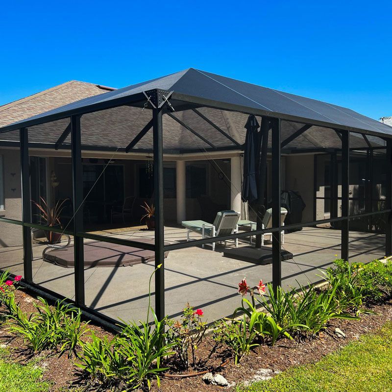 Screened patio enclosure with black frame and dark roof beside a house, with chairs and garden plants