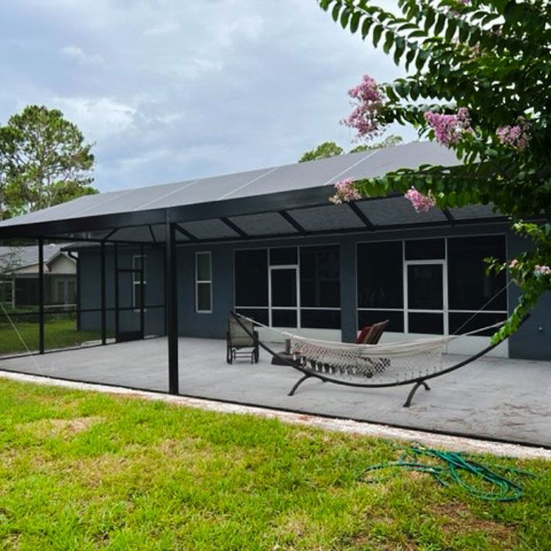 Dark gray house with a metal awning, patio chairs, hammock, and green lawn under cloudy sky