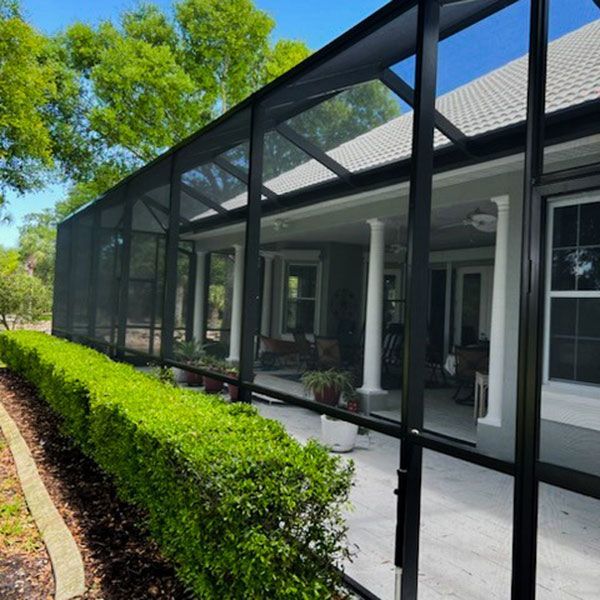 Screened patio beside a house, with black framing, patio furniture, and neatly trimmed hedges along the walkway.