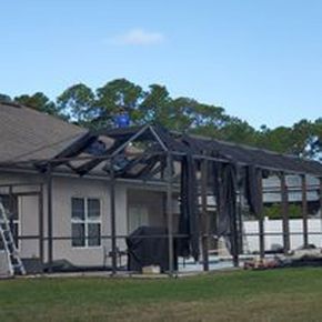 Damaged screened porch roof and frame beside a house, with a ladder and green lawn.