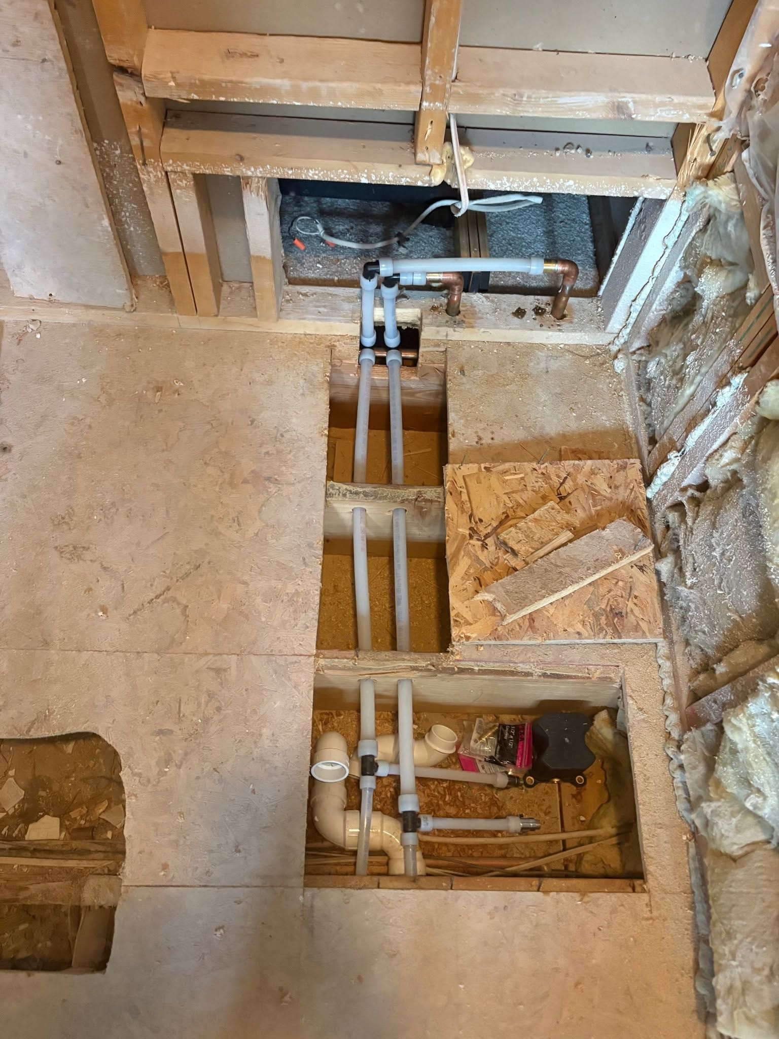 Open floor with exposed plumbing pipes, joists, and insulation during home renovation.
