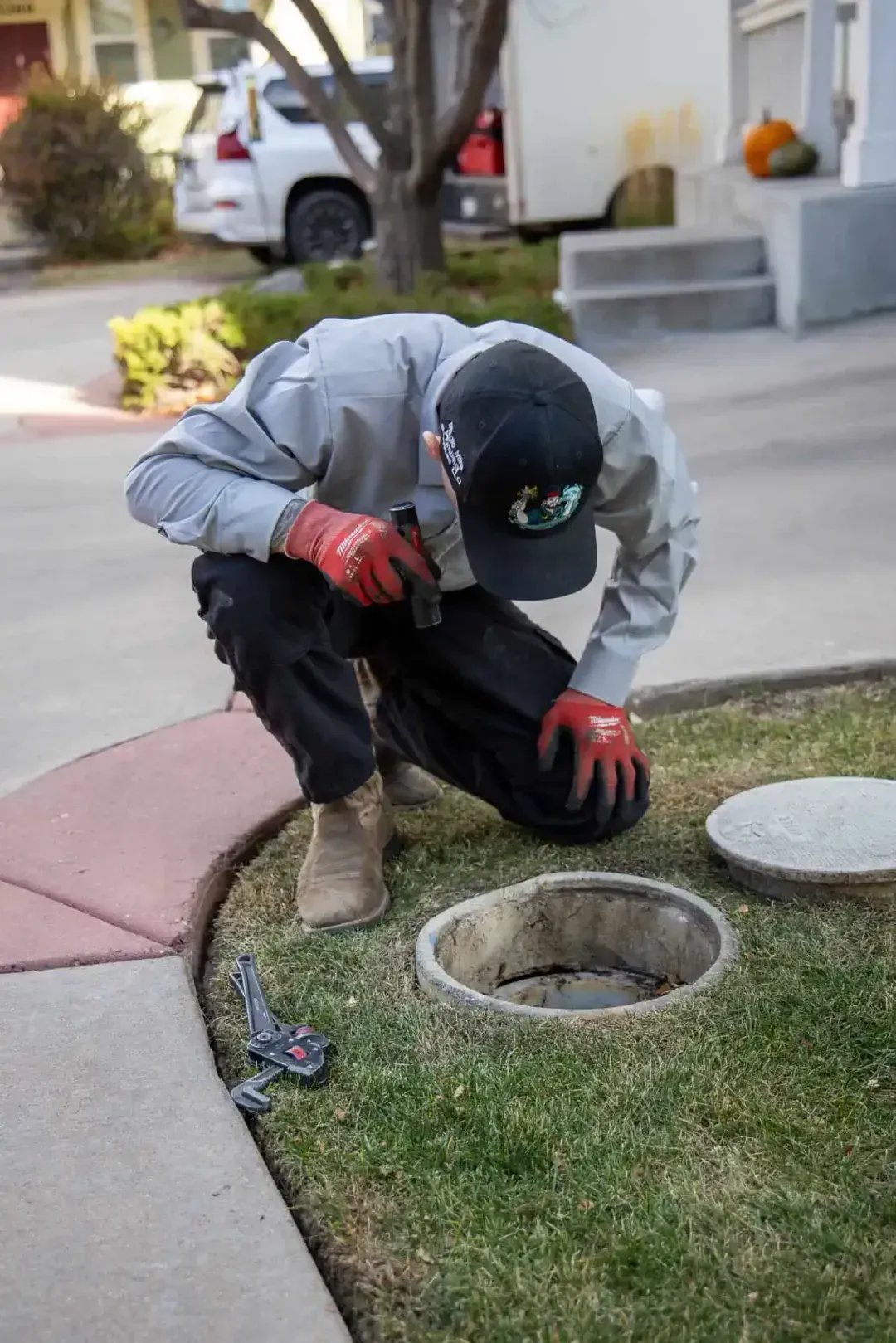 Worker kneeling by a lawn sprinkler valve box in a suburban yard, inspecting it with gloved hands.