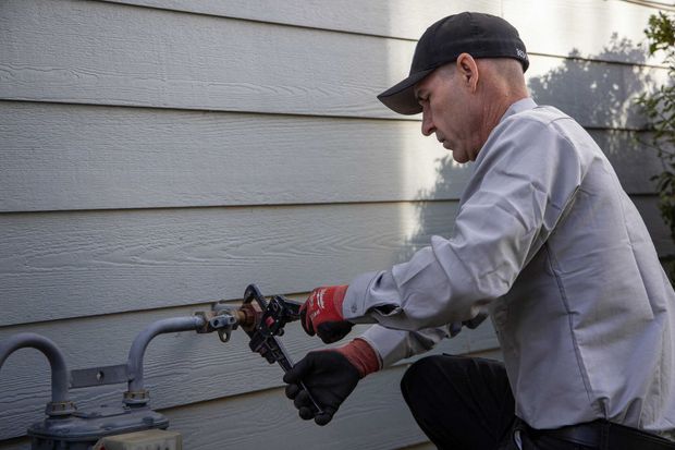 Worker in gray shirt and black cap using a tool on a gas pipe beside a house wall