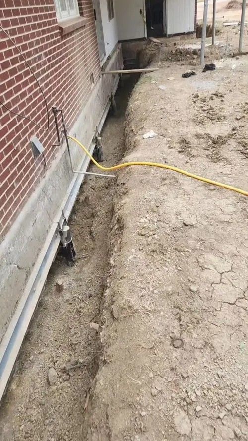 Narrow dirt trench beside a brick house with a yellow hose stretched across it and a concrete edge on the left