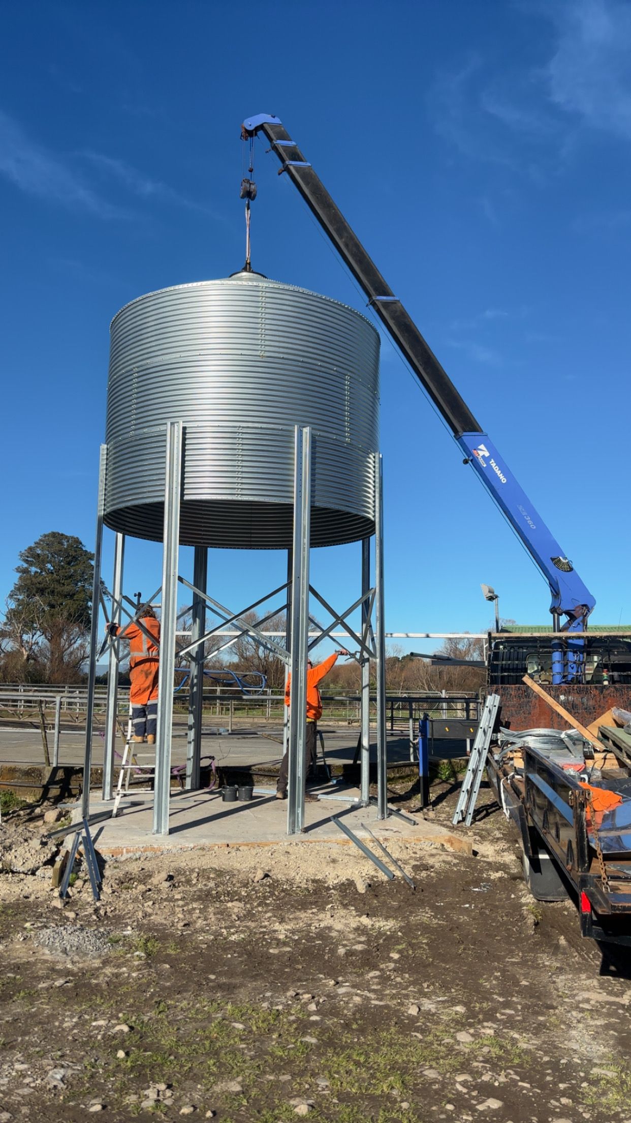 Crane lifting a large silver water tank on steel supports at a construction site under a blue sky