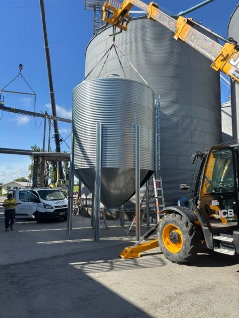 A grain silo being lifted into place by a crane. Construction site with workers, a forklift, and other silos.