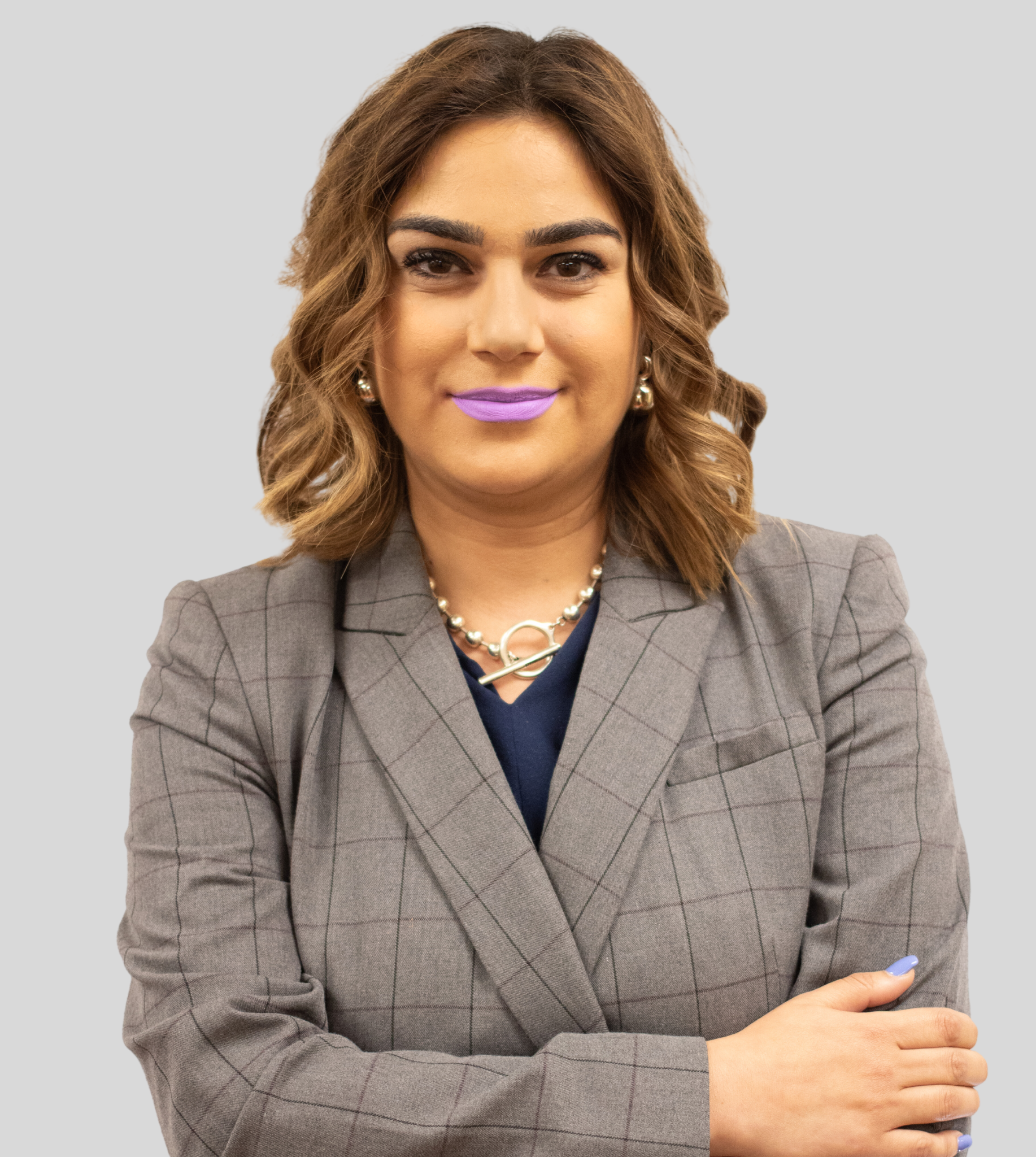 A person with shoulder-length wavy hair wearing a grey plaid blazer and a silver chain necklace, standing against a grey.