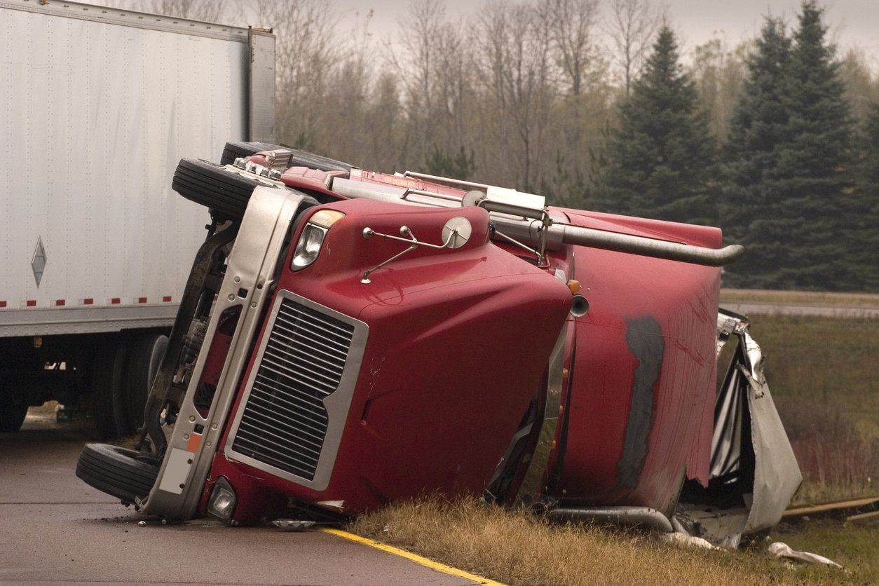 A red semi-truck lies on its side on the grassy shoulder next to a road, with its trailer attached.