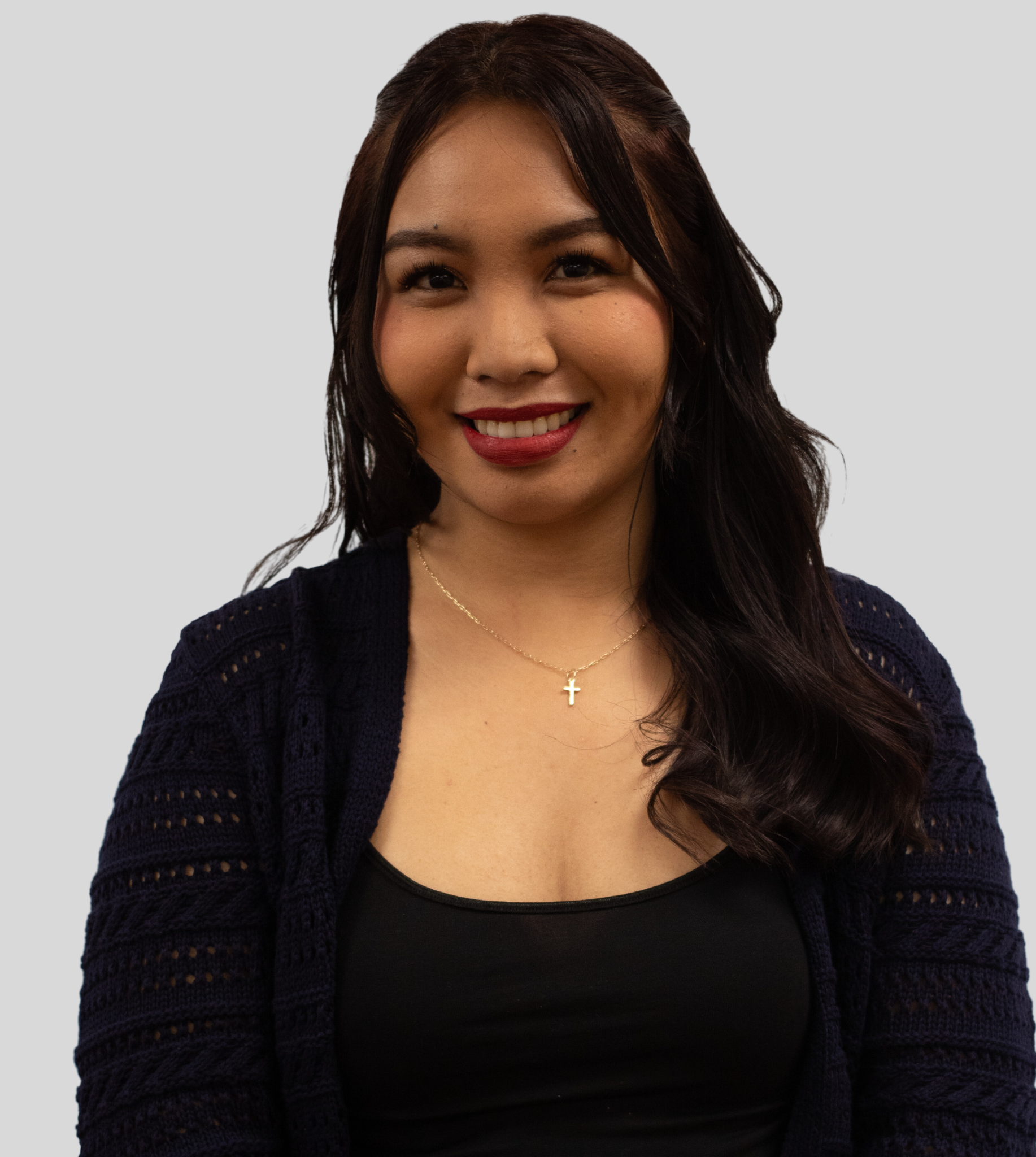 A smiling person with long, dark, wavy hair, wearing a dark blue knit cardigan over a black top and a small cross necklace.
