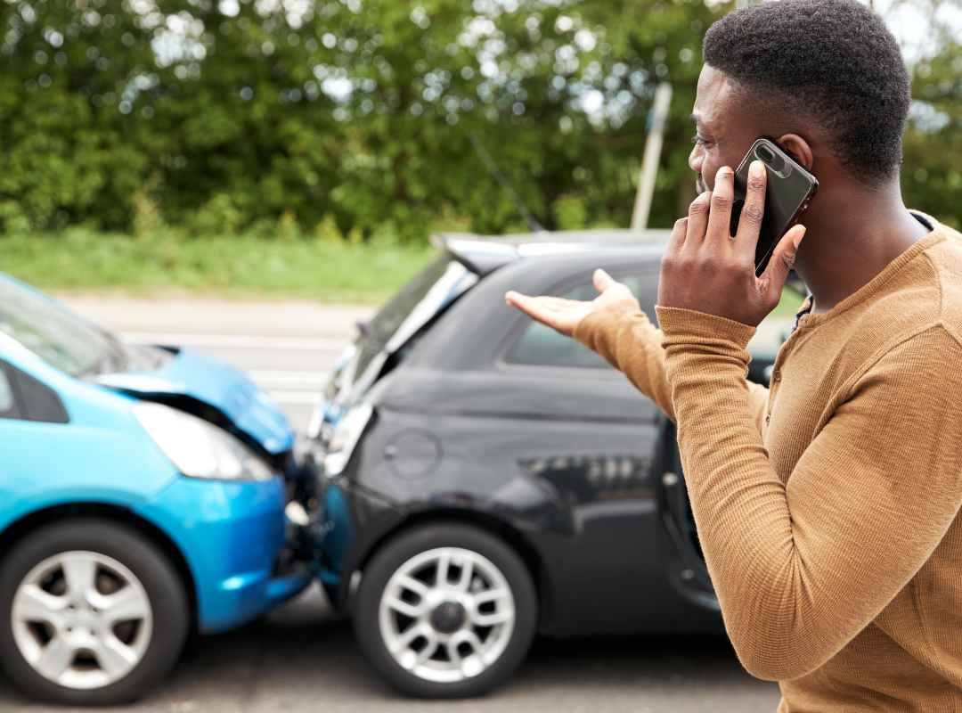 A person in a brown shirt talks on a phone while gesturing toward a blue car that has rear-ended a black car.