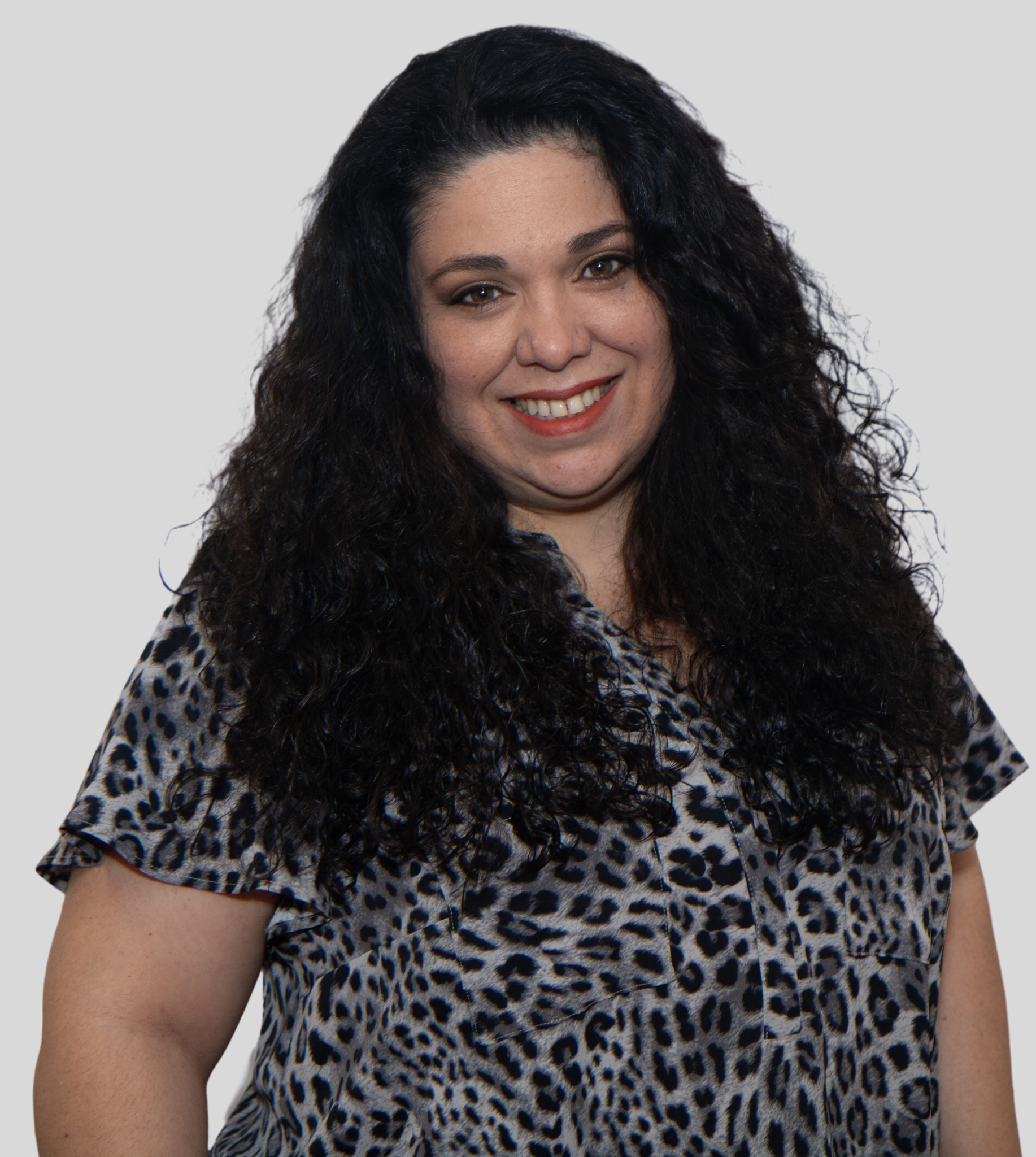 A person with long, curly dark hair smiles at the camera, wearing a grey and black leopard-print short-sleeved top.