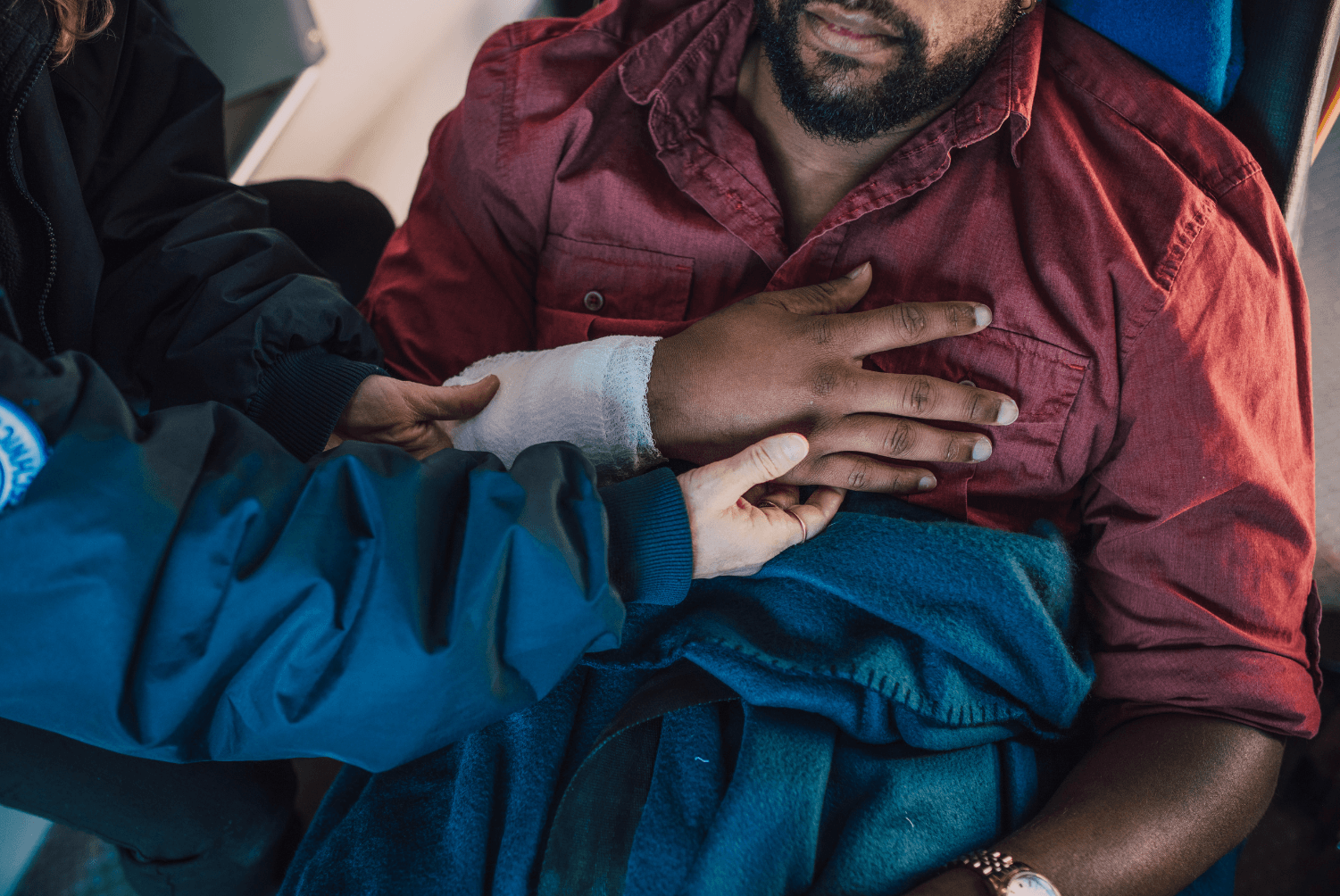 A medical professional in a blue jacket examines a person's bandaged hand resting on their chest.