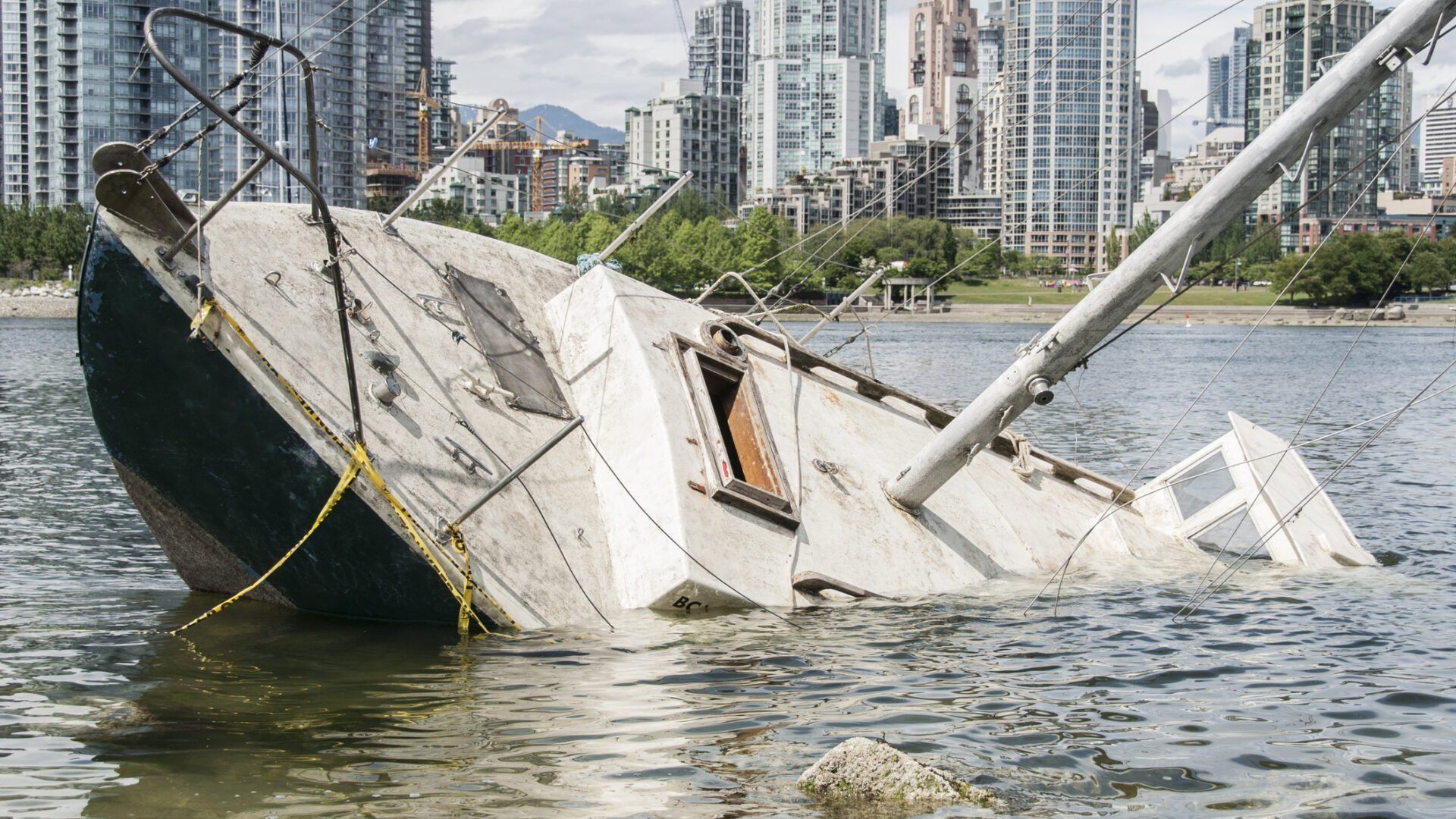 A partially submerged sailboat resting on its side in shallow water, with a cityscape of tall buildings in the background.