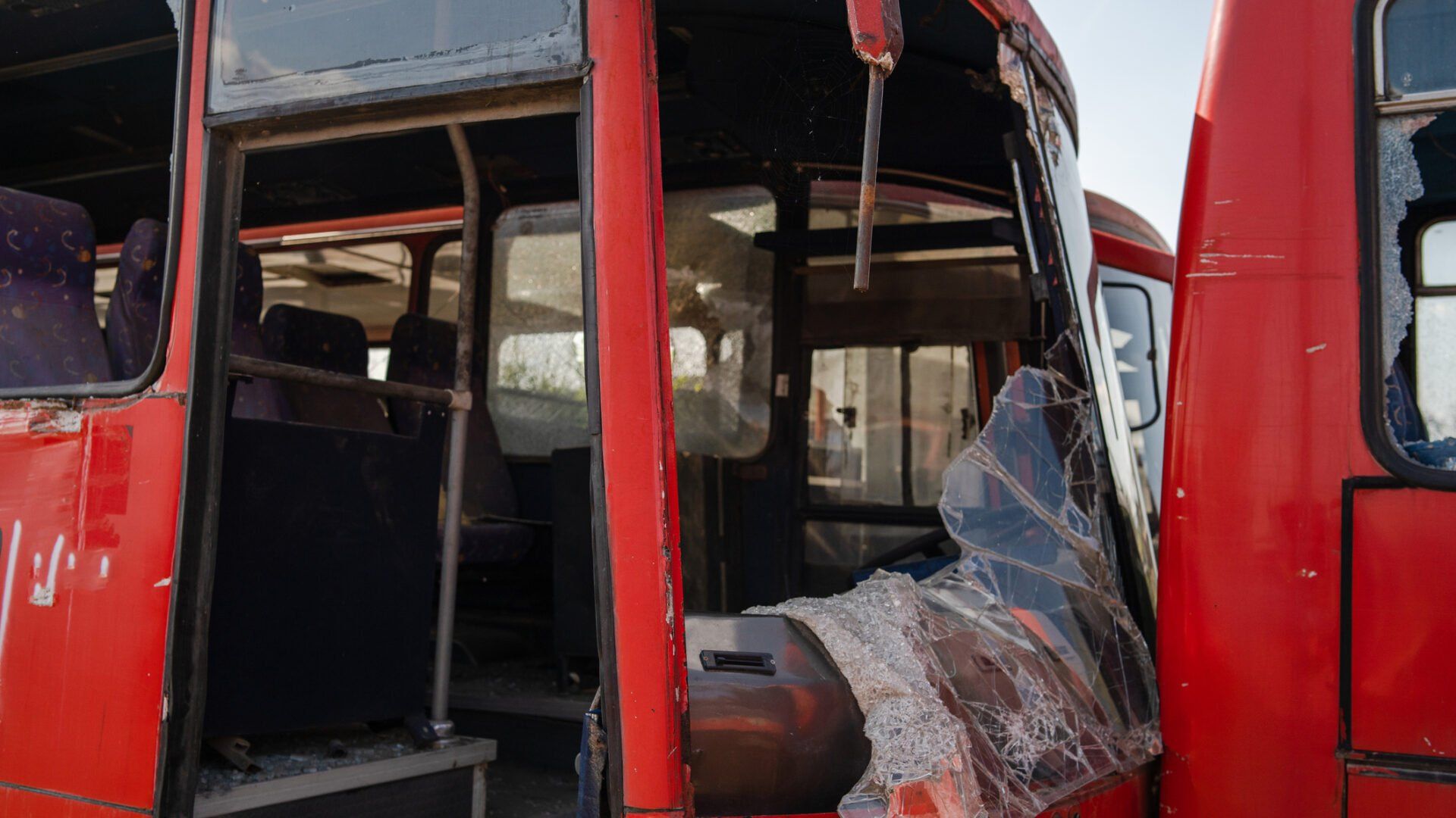 A red bus with a shattered windshield and heavily damaged exterior frame, showing empty seats inside.