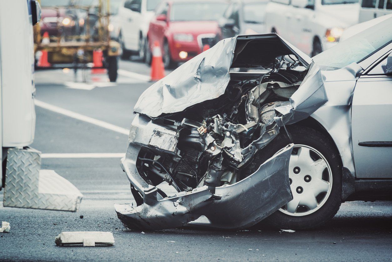 A silver car with extensive front-end damage after a collision, parked on a road with other vehicles in the background.