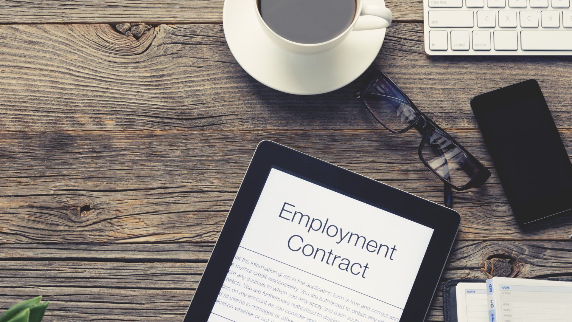 A tablet showing an employment contract on a rustic wooden desk with a coffee cup, glasses, keyboard, and smartphone.