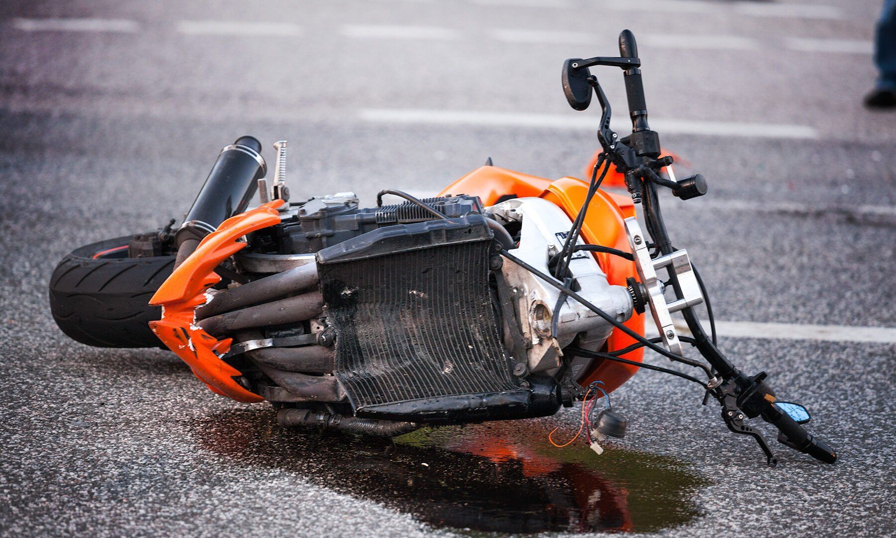 An orange motorcycle lying on its side on the asphalt, with a small puddle of leaked fluid beneath the radiator.
